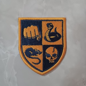 May include: A yellow and blue embroidered patch with four different symbols: a fist, a snake, a rat, and a skull. The patch is shaped like a shield.
