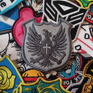 Puede incluir: Un parche bordado gris con un águila negra sosteniendo una cruz. El águila tiene las alas extendidas y está posada sobre un escudo.