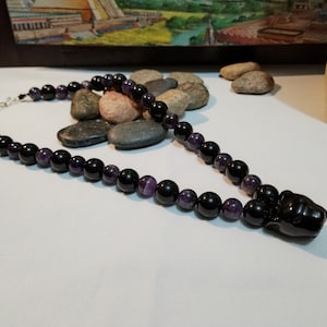 May include: A beaded necklace featuring alternating black and purple beads. The necklace has a silver clasp and a black panther pendant. The beads are round and smooth, creating a striking contrast.