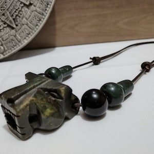 May include: A dark green stone necklace featuring a carved jaguar head pendant. The necklace includes dark green and black beads strung on a brown leather cord. The jaguar head is intricately carved with detailed features.