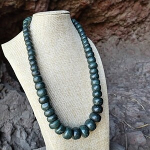 May include: A dark green beaded necklace displayed on a beige bust. The beads are round and vary in size, creating a graduated effect. The necklace is made of a dark green material and is set against a natural, textured background.