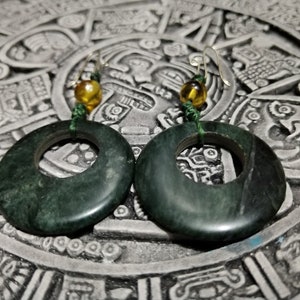 Puede incluir: Un par de pendientes de jade verde oscuro. Cada pendiente presenta un colgante circular grande y pulido de jade con un agujero en el centro. Los pendientes están suspendidos de ganchos dorados con una pequeña cuenta de color ámbar y una cuerda verde.