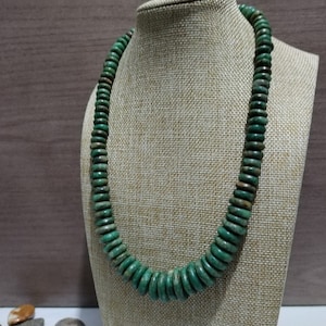 Puede incluir: Un collar de cuentas verdes expuesto en un maniquí. El collar está compuesto por numerosas cuentas redondas y planas en varios tonos de verde. Las cuentas están ensartadas para formar una pieza de joyería llamativa. Un accesorio llamativo.