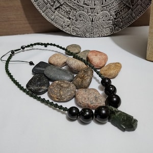 Puede incluir: Un collar hecho a mano con un cordón de macramé verde oscuro, cuentas negras y un colgante verde tallado. El collar se exhibe sobre una superficie blanca, con una colección de piedras en el fondo.