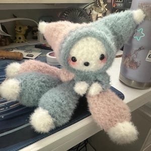 May include: A plush toy featuring a fuzzy, multi-colored design in pastel blue, pink, and white. The creature has a hood, round black eyes, and pink cheeks. It has long, soft limbs with white tips, a rounded body, and a textured, yarn-like appearance.