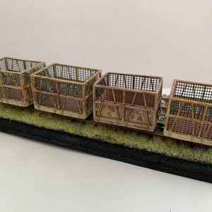 Può includere: Un modello in miniatura di quattro vagoni ferroviari in metallo arrugginito e usurato, con lati in rete metallica. I vagoni poggiano su un binario in miniatura con erba verde e sono montati su una base nera. Il modello ha un aspetto vintage e invecchiato.