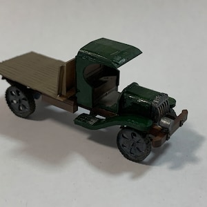 May include: A green toy truck with a brown wooden bed. The truck has a cab with an open hood and a detailed engine. The truck is on a white surface.
