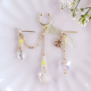 May include: A set of three gold-toned earrings with delicate charms. The earrings feature a butterfly, a leaf, and a sun charm. The charms are made of clear and yellow glass beads.