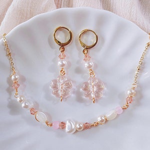 Pode incluir: Um conjunto delicado de colar e brincos de ouro com contas de vidro rosa e branco e pingentes em forma de flor.