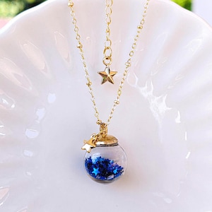 May include: A gold-coloured necklace with a clear glass orb pendant filled with blue star-shaped glitter. The pendant has a gold-coloured cap and a small star charm. The necklace chain is delicate.