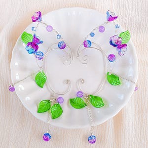 May include: A pair of silver ear cuffs with a floral and leaf design. The cuffs feature translucent green leaves, purple and blue flowers, and purple and blue beads. The ear cuffs are displayed on a white plate.