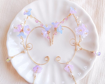 May include: Elegant gold-coloured ear cuffs featuring pastel purple and blue flower details, tiny pearls, and dangling crystal charms. The floral design is arranged on a curved wire frame, creating a whimsical aesthetic. Ideal for special events and celebrations.