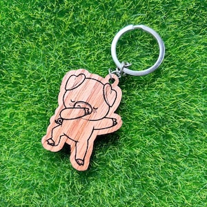 Könnte beinhalten: Holzschlüsselanhänger mit einem Cartoon-Schwein, das ein herzförmiges Stirnband trägt und den Dab macht. Der Schlüsselanhänger ist an einem silbernen Metallring befestigt.