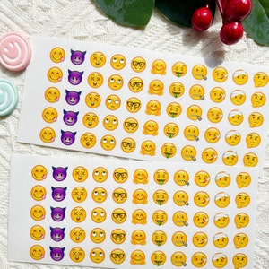 May include: A sheet of yellow and purple emoji stickers with various expressions, including winking, tongue sticking out, devil horns, and money bags. The stickers are arranged in rows on a white background.
