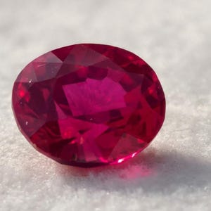 May include: A faceted oval-shaped gemstone with a deep, vibrant red colour. The stone features numerous triangular and trapezoidal facets that reflect light, creating a brilliant, sparkling appearance across its polished, translucent surface.