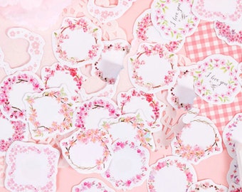May include: A set of pink and white floral decorative stickers with a blank space in the center for writing. The stickers feature a delicate floral design with a pink and white color scheme.