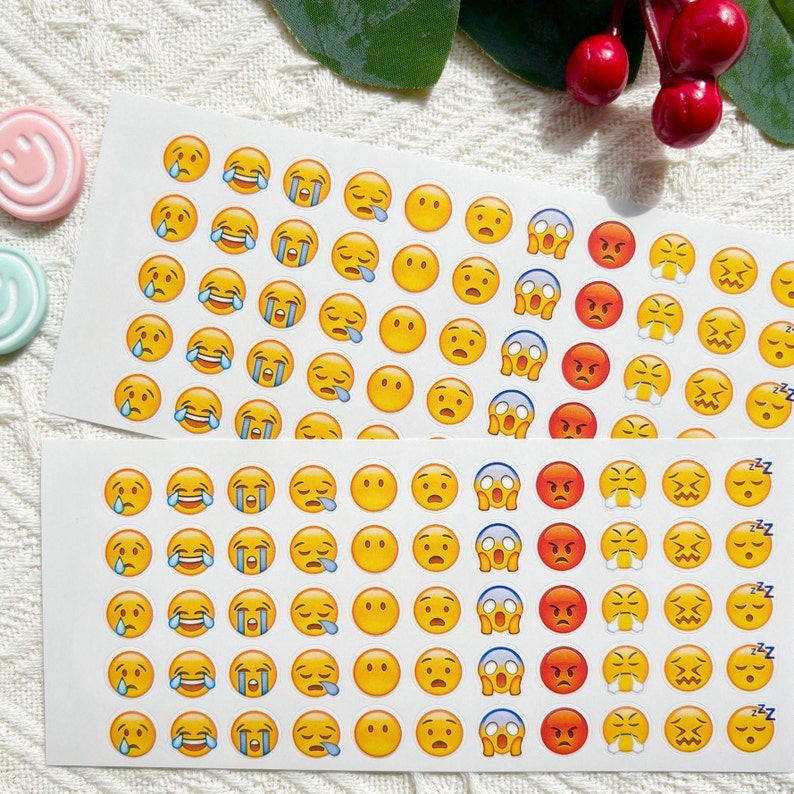 May include: Two sheets of white paper with yellow and blue emoji stickers. The emojis include smiling faces, crying faces, surprised faces, and sleeping faces.