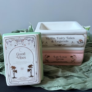 Könnte beinhalten: Ein Keramik-Wachswärmer in Form eines Stapels aus zwei Büchern. Das obere Buch ist weiß mit dem Text Myths, Fairy Tales & Legends. Das untere Buch ist rosa mit dem Text Let Love Grow. Ein separater weißer Deckel zeigt den Text Good Vibes.