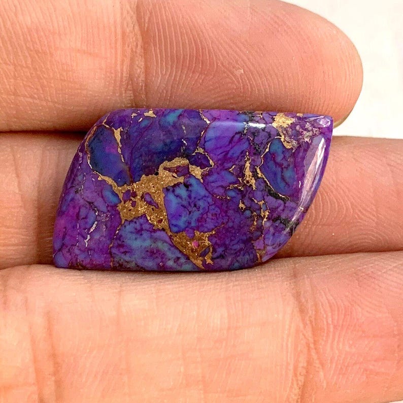 May include: A polished, gemstone with a vibrant purple base, interspersed with teal and gold veining. The stone is held in a hand, showcasing its unique color and pattern. The shape is a rounded rectangle.