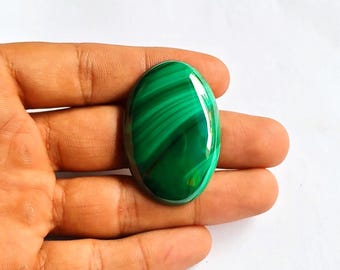 May include: Oval-shaped malachite gemstone with a polished, glossy surface. The stone features a vibrant green color with darker green, swirling patterns. The gemstone is held in a hand against a white background. This gemstone is suitable for jewelry making.