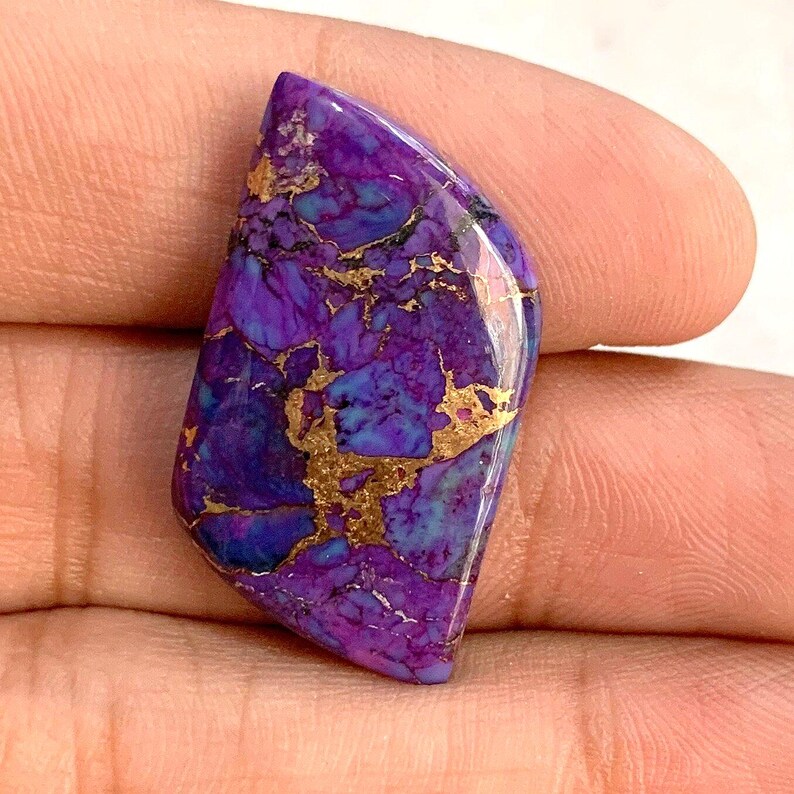 May include: A polished gemstone with a vibrant color palette. The stone features a mix of deep purple, blue, and gold hues. The gold veins create a striking contrast against the purple and blue base, giving it a unique appearance. The stone is held in a hand.
