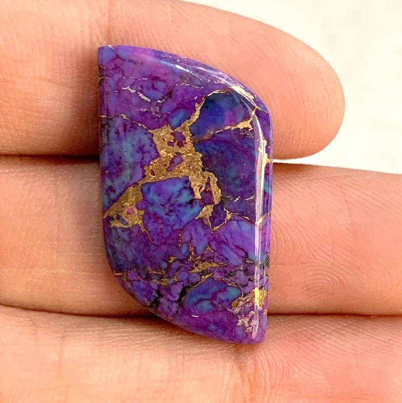 May include: A polished gemstone with a vibrant mix of purple, blue, and gold colors. The stone has a unique pattern with gold veins running through the purple and blue base. The shape is a rounded rectangle, perfect for jewelry making.