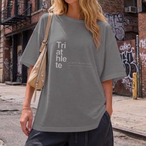 May include: Gray oversized t-shirt with the word "Triathlete" printed vertically on the front. The shirt has short sleeves and a relaxed fit. The text "I AM THE SURFING..." is printed below the word "Triathlete".