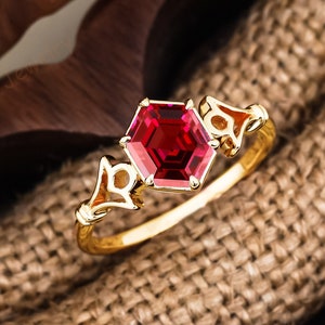 May include: A gold ring with a red gemstone in a hexagonal shape. The ring is set in a delicate, ornate design with intricate details. The ring is displayed in a wooden box lined with burlap.