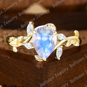 May include: A gold ring featuring a pear-shaped, iridescent gemstone, accented with small, clear stones and leaf-like details. The ring is set against a dark wooden background, showcasing the jewelry's design.