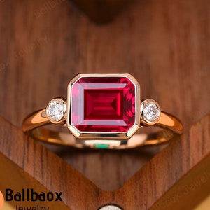 May include: A rose gold ring featuring a large, emerald-cut ruby gemstone with two smaller round diamonds on either side.