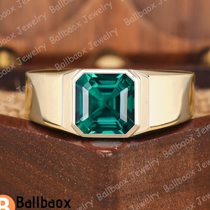Könnte beinhalten: Ein goldfarbener Ring mit einem großen, smaragdgeschliffenen, tiefgrünen Edelstein in der Mitte. Der Ring hat ein dickes Band und eine rechteckige Fassung, die die Form des Edelsteins ergänzt. Der Ring wird auf einer Holzoberfläche präsentiert.