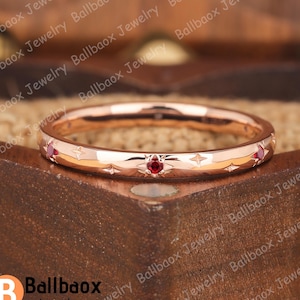 May include: Rose gold ring with a repeating pattern of starbursts and small red gemstones. The ring is displayed on a wooden surface. The ring is from Ballbaox Jewelry.