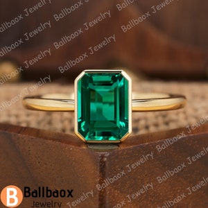 May include: A gold ring with a large emerald green gemstone set in a square shape. The ring is on a wooden surface.