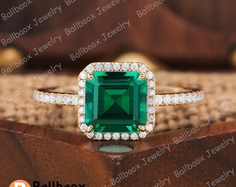 May include: A gold ring featuring a square emerald green gemstone surrounded by a halo of smaller diamonds. The band is also set with smaller diamonds.