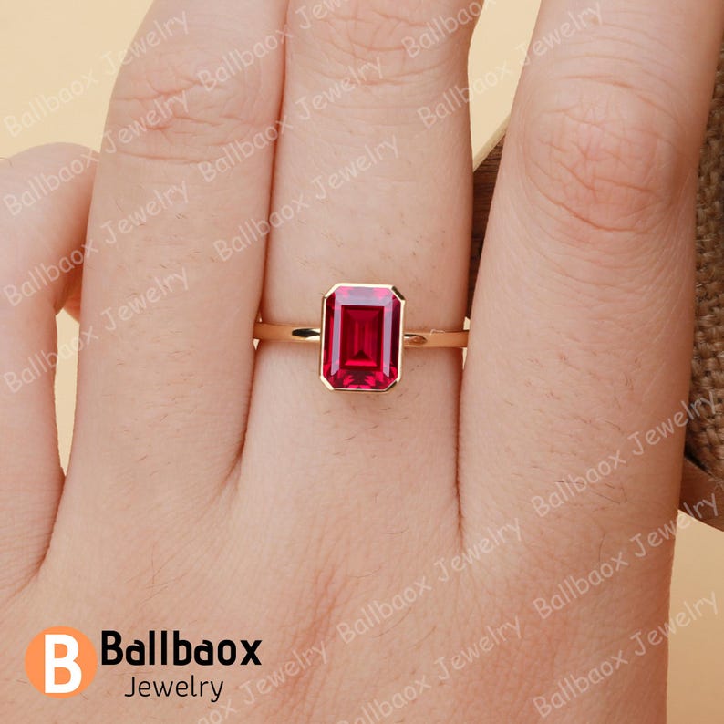 May include: A gold ring featuring a rectangular, deep red gemstone. The ring's band is thin and gold-colored, with the gemstone set in a bezel setting. The ring is displayed on a hand, showcasing the jewelry's design.