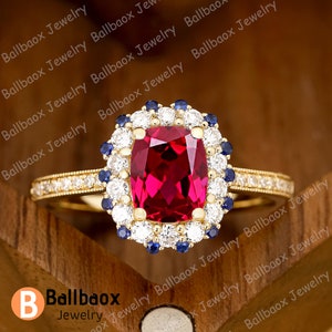 Puede incluir: Un anillo de oro con una gran piedra preciosa roja rodeada de un halo de piedras preciosas blancas y azules más pequeñas.