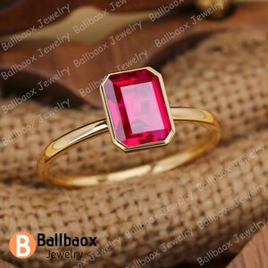 May include: A gold ring featuring a large, rectangular, ruby-red gemstone. The ring's band is a polished gold, and the gemstone is set in a matching gold bezel. The ring is displayed on a textured, neutral-toned surface.