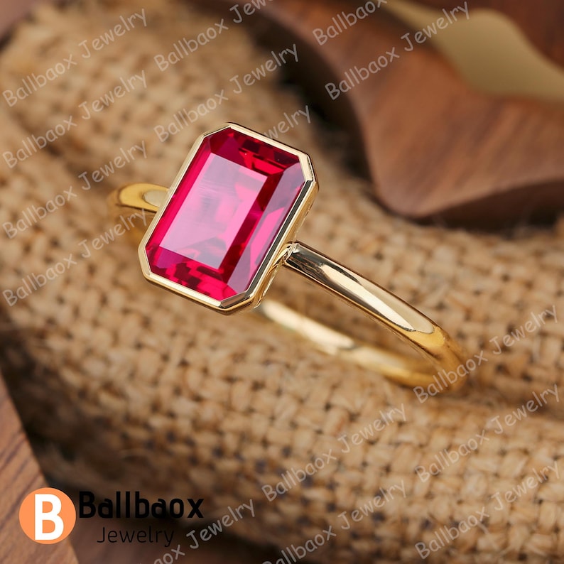 May include: A gold ring featuring a rectangular, vibrant red gemstone. The ring has a simple, elegant design with a polished gold band. The gemstone is set in a gold bezel, enhancing its color and shape. The ring is a piece of jewelry.
