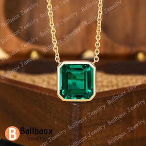May include: A gold necklace featuring a square, emerald-cut green gemstone pendant. The pendant is set in a gold bezel and suspended from a delicate gold chain. The necklace is presented on a wooden surface.