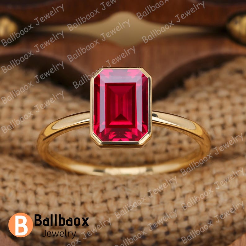 May include: A gold ring featuring a rectangular, vibrant red gemstone. The ring has a simple, elegant design with a thin band. The gemstone is set in a bezel setting, enhancing its color and shape. The ring is a piece of jewelry.