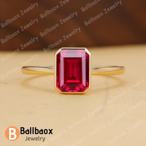 May include: A gold ring featuring a rectangular, deep red gemstone. The gemstone is set in a gold bezel, and the band is a simple, polished gold. The ring is displayed on a neutral background. The brand name "Ballbaox Jewelry" is visible.