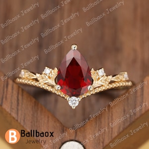 Puede incluir: Un anillo de oro con una piedra roja en forma de pera y un delicado diseño floral. El anillo está engastado en una banda de oro con una fila de pequeños diamantes a cada lado de la piedra principal.