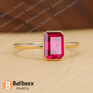 May include: A gold ring featuring a rectangular, vibrant red gemstone. The gemstone is set in a gold bezel, and the band is a simple, polished gold. The ring is displayed on a neutral background, highlighting the jewelry's design.