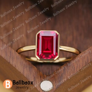 May include: A gold ring featuring a large, rectangular, vibrant red gemstone. The gemstone is set in a bezel setting. The ring is displayed on a wooden surface. The brand name "Ballbaox Jewelry" is visible.