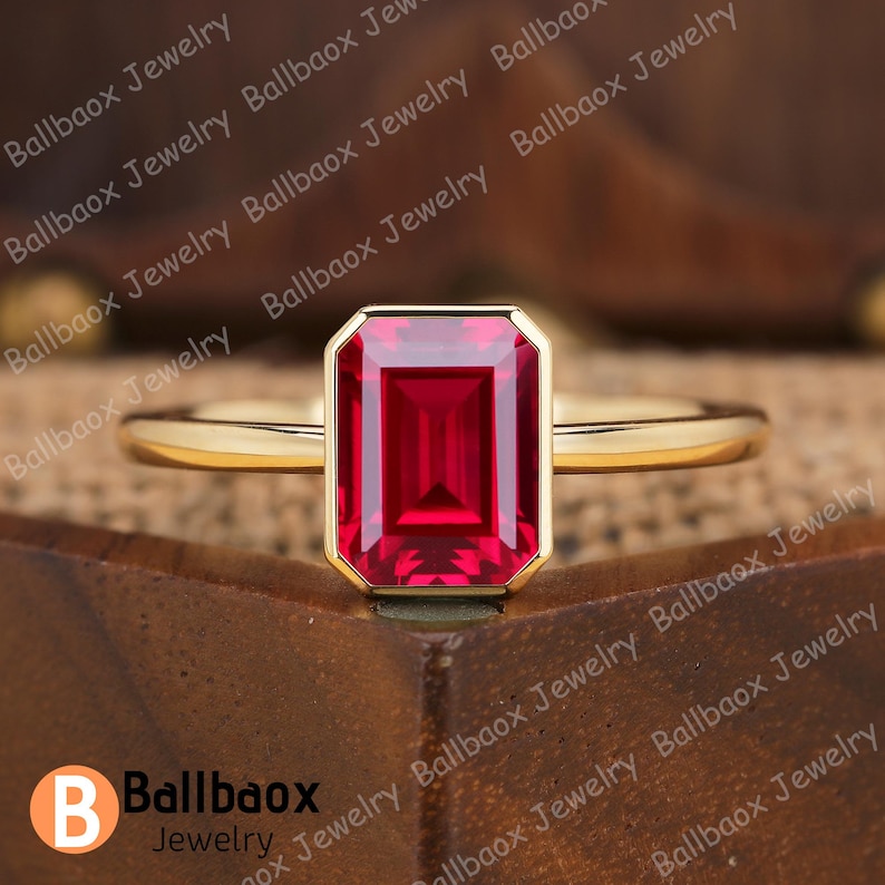May include: A gold ring featuring a rectangular, deep red gemstone. The gemstone is set within a gold bezel, and the band is a simple, polished gold. The ring is displayed on a wooden surface. The brand name "Ballbaox Jewelry" is visible.