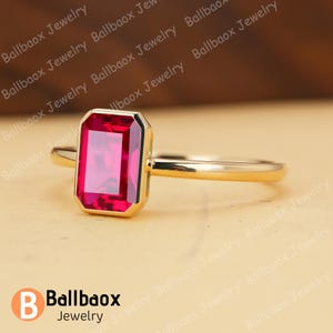 May include: A gold ring featuring a rectangular, vibrant red gemstone. The gemstone is set within a gold bezel, and the band is a simple, polished gold. The ring is displayed on a neutral background. The brand name "Ballbaox Jewelry" is visible.