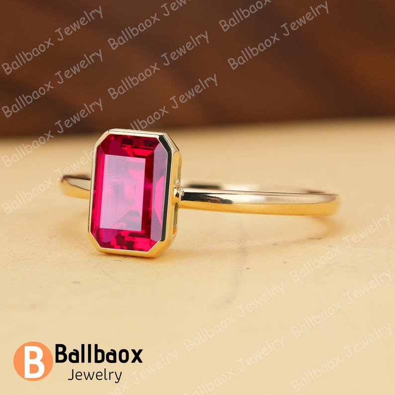 May include: A gold ring featuring a rectangular, vibrant red gemstone. The gemstone is set within a gold bezel, and the band is a simple, polished gold. The ring is displayed on a neutral background. The brand name "Ballbaox Jewelry" is visible.