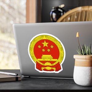 May include: A sticker featuring the emblem of the People's Republic of China. The emblem is a red circle with a gold border, containing five gold stars and a depiction of Tiananmen Square. Red ribbons and wheat stalks complete the design.