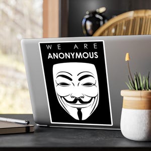 Peut inclure: Un autocollant noir et blanc avec le masque de Guy Fawkes et le texte « WE ARE ANONYMOUS ». L'autocollant rectangulaire est collé sur un ordinateur portable. Le masque affiche un sourire et une moustache stylisés.