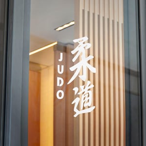 May include: A glass door with the word "JUDO" in white block letters on the left and Japanese characters for "Judo" on the right. The background features vertical wooden slats.